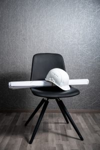 Bauhelm und Pläne auf schwarzem Stuhl vor strukturierter Wand, Symbol für Architektur.